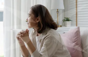 Woman seated by a bright window, hands clasped in thought, with a pink cushion and plant in a cozy living room setting.