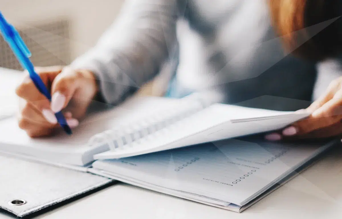 Person writes with a blue pen on papers at a desk, with a notebook nearby and a tablet nearby.