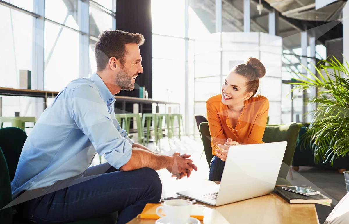 Two coworkers converse in a bright modern office; a bearded man in a blue shirt talks with a smiling woman in an orange blouse across a laptop on the table.