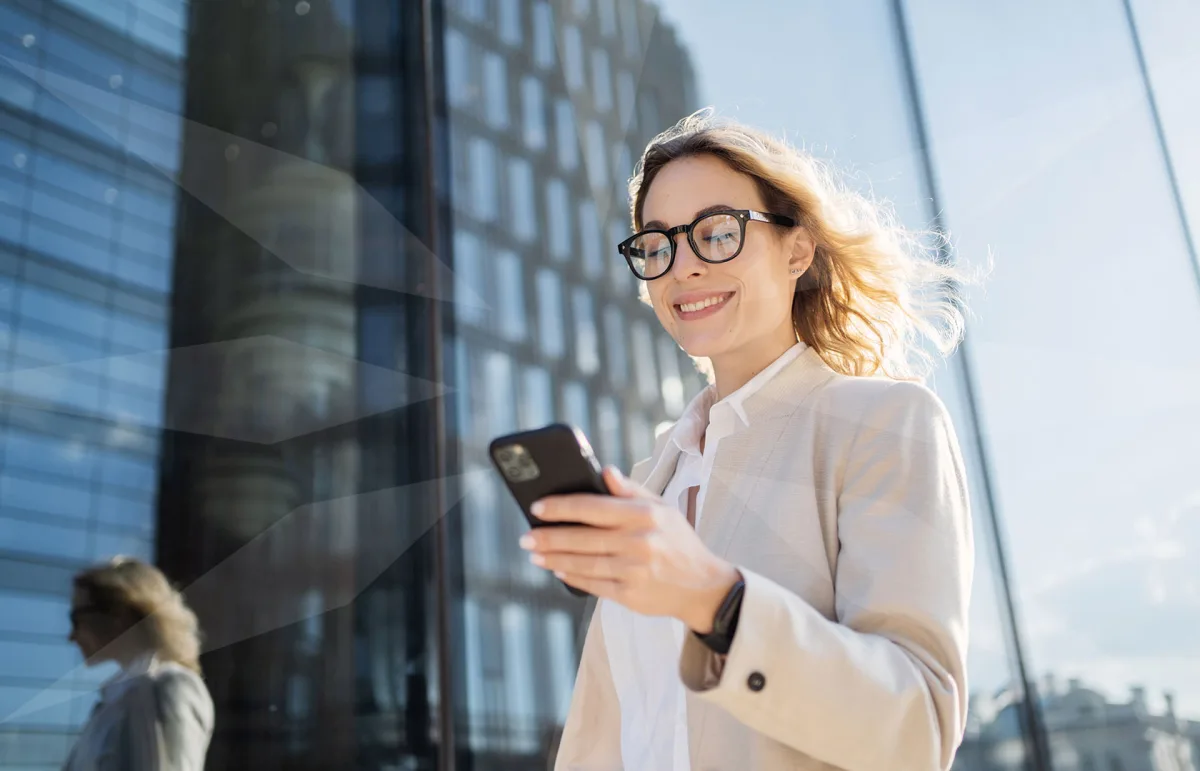 Businesswoman wearing a beige blazer and glasses, smiling as she uses a smartphone outside a glass office building.