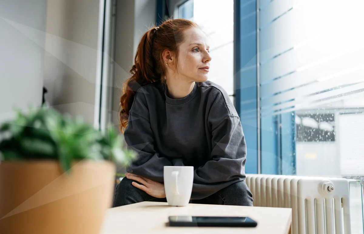 Side-profile of a woman with red hair in a ponytail, sitting by a window with a white mug on the table, looking out thoughtfully.