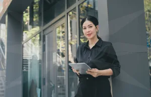 Professional woman in black outfit standing outside a modern glass building, holding a tablet and looking at the camera.