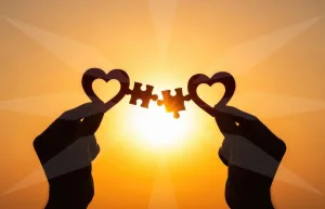 Two silhouetted hands hold heart-shaped cutouts with interlocking puzzle pieces against a bright sunrise.