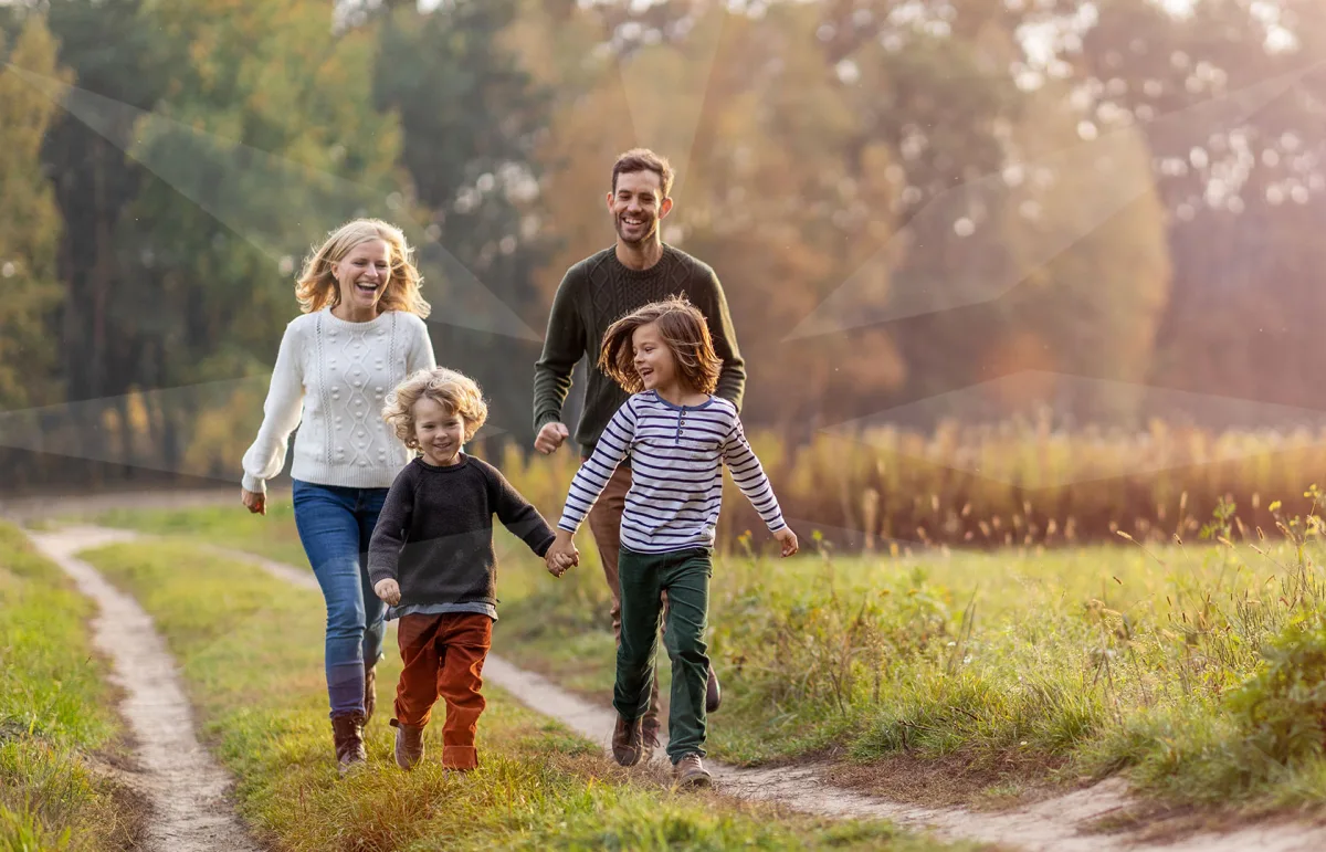 A family of five walking along a dirt path in a sunny park, holding hands and laughing together.
