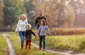 A family of five walking along a dirt path in a sunny park, holding hands and laughing together.