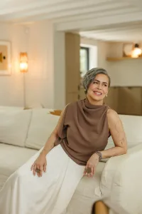 Smiling older woman with short gray hair sits on a white sofa in a bright, modern living room, wearing a taupe sleeveless top and white skirt.
