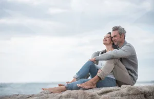 Couple sitting on rocks by the ocean, arms around each other, smiling at the sea under a cloudy sky.